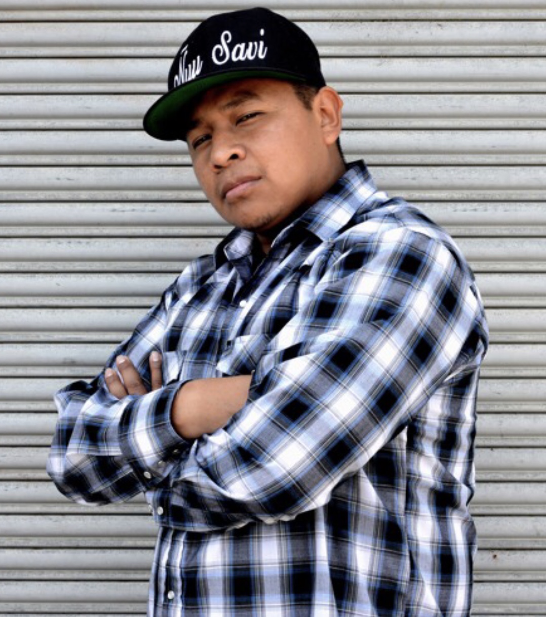 man with his arms crossed looking at the camera intently and has a snapback on